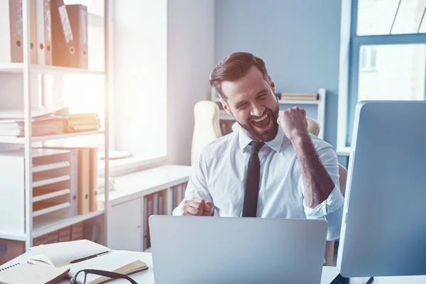 Vrolijke jonge man in shirt en das werken met behulp van de computer en glimlachen tijdens het zitten in het kantoor — Stockfoto