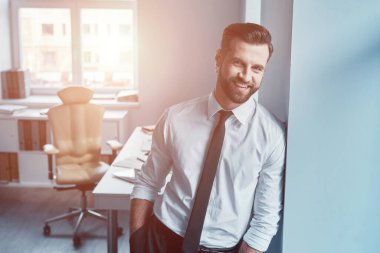 Gömlekli ve kravatlı zarif bir genç. Ofiste dururken kameraya bakıyor ve gülümsüyor.