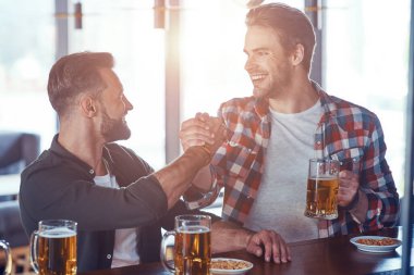 Barda bira içerken el sıkışan ve gülümseyen iki genç adam.