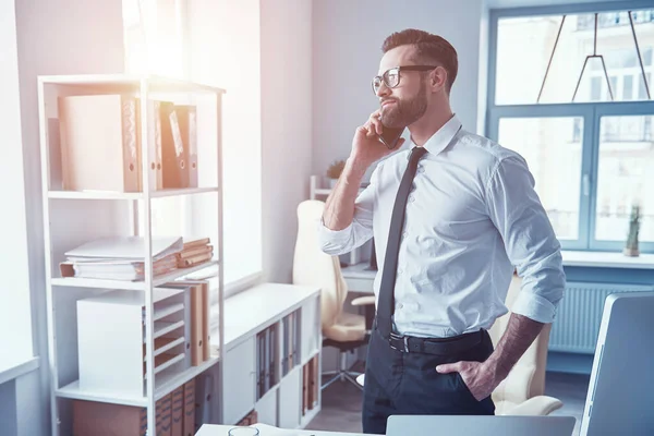 Hezký mladý muž ve formálním oděvu mluví do telefonu a usmívá se při práci v kanceláři — Stock fotografie