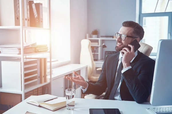 Hombre joven guapo en camisa y corbata hablando en el teléfono inteligente y sonriendo mientras está sentado en la oficina — Foto de Stock