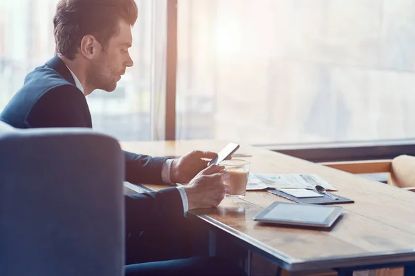 Side view of handsome young businessman in full suit using smart phone and drinking coffee — Stock Photo, Image