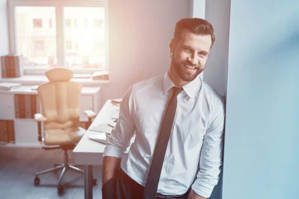 Elegante jongeman in hemd en das die naar de camera kijkt en glimlacht terwijl hij op kantoor staat — Stockfoto