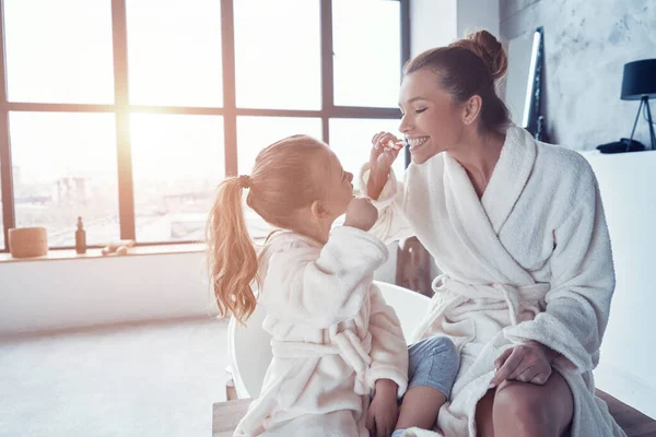 Madre e hija en albornoces sonriendo y limpiando los dientes mientras hacen la rutina de la mañana — Foto de Stock