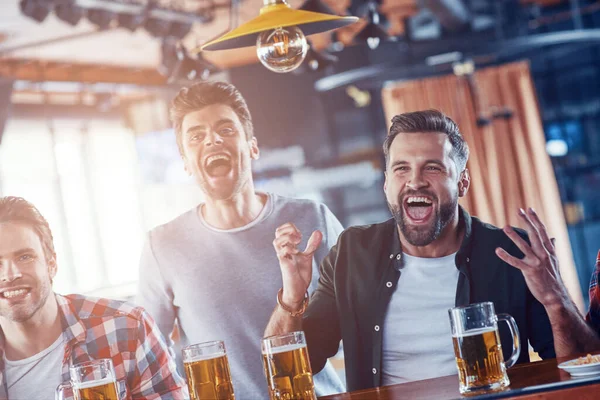 Vrolijke jonge mannen in casual kleding drinken bier en kijken naar sport wedstrijd tijdens het zitten in de pub — Stockfoto