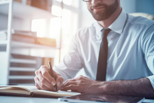 Close up of young man in formalwear writing something down while sitting in the office — Stock Photo, Image