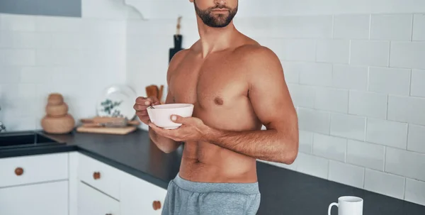 Hombre joven sin camisa de buen aspecto desayunando mientras está de pie en la cocina casera — Foto de Stock