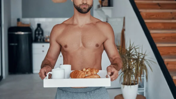 Bello giovane uomo senza maglietta che tiene il vassoio con la colazione mentre trascorre del tempo a casa — Foto Stock