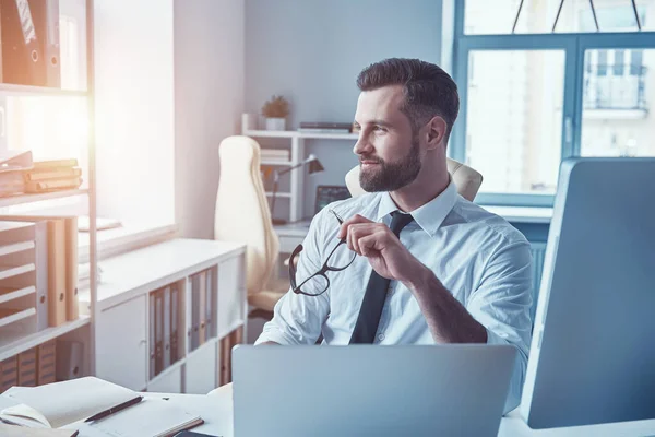 Jeune homme réfléchi en tenue de cérémonie regardant ailleurs et souriant assis dans le bureau — Photo