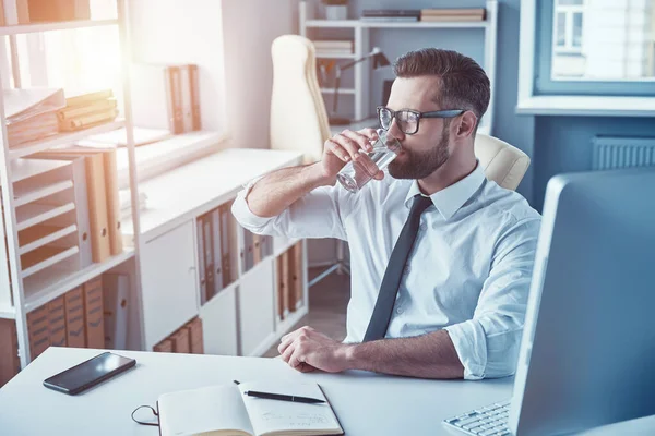Dobře vypadající mladý muž v košili a kravatu pitnou vodu, zatímco sedí v kanceláři — Stock fotografie
