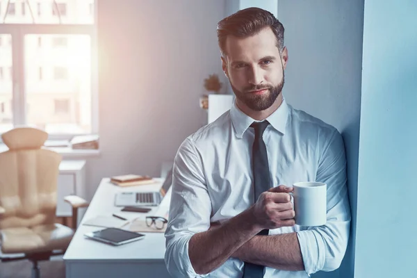 Goed uitziende jonge man in shirt en stropdas holding cup en glimlachen terwijl staan in het kantoor — Stockfoto