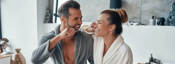 Sabah rutinini yaparken gülümseyen ve dişlerini temizleyen bornozlu genç çift. — Stok fotoğraf
