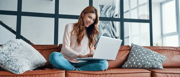 Mujer joven atractiva usando el ordenador portátil y sonriendo mientras pasa tiempo en casa —  Fotos de Stock