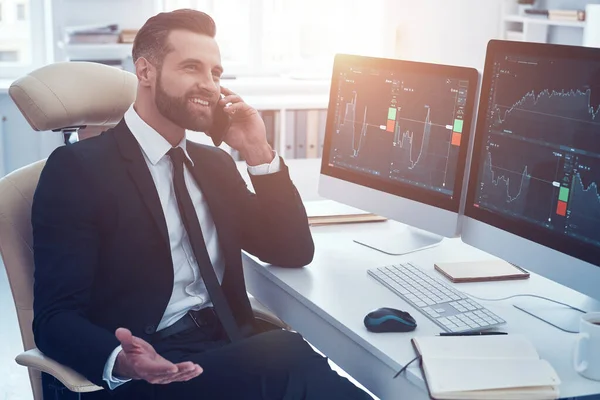 Усміхнений молодий чоловік в сорочці і краватці аналізує дані на фондовому ринку і розмовляє по телефону під час роботи в офісі — стокове фото
