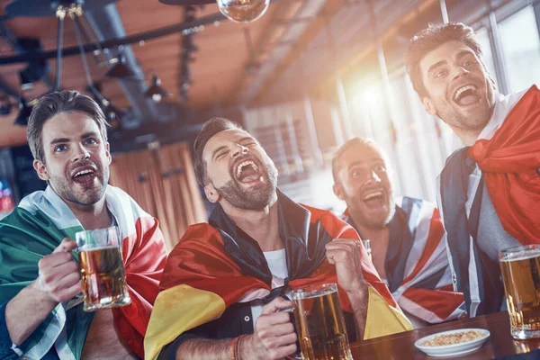 Jóvenes felices cubiertos de banderas internacionales bebiendo cerveza y viendo el deporte mientras están sentados en el pub —  Fotos de Stock