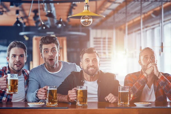 Vrolijke jonge mannen in casual kleding drinken bier en kijken naar sport wedstrijd tijdens het zitten in de pub — Stockfoto