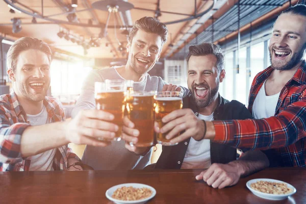 Happy young men in casual clothing toasting each other with beer and laughing while sitting in the pub — Stock Photo, Image