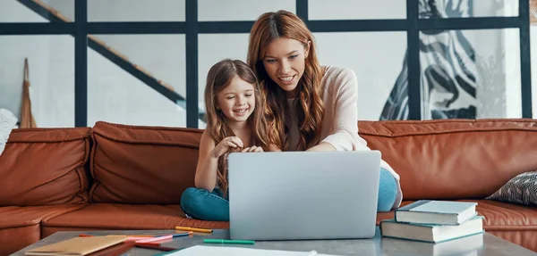 Fiatal gyönyörű anya és kislánya összebarátkoznak és mosolyognak, miközben otthon laptopot használnak. — Stock Fotó