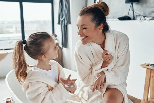 Madre con hija en albornoces sonriendo y usando crema de manos mientras hace la rutina de la mañana —  Fotos de Stock