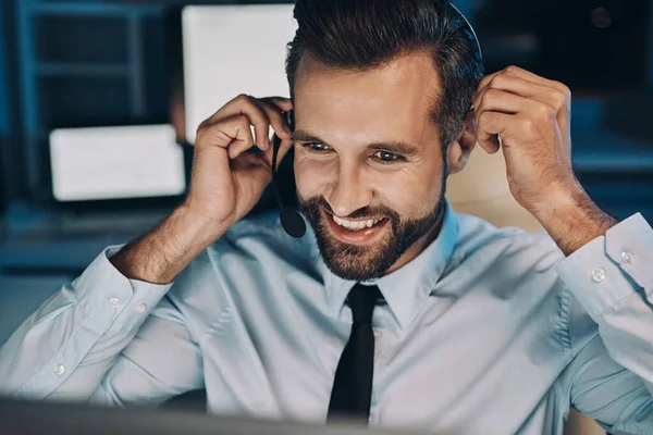 Jovem feliz no fone de ouvido olhando para o computador e sorrindo enquanto fica até tarde no escritório — Fotografia de Stock