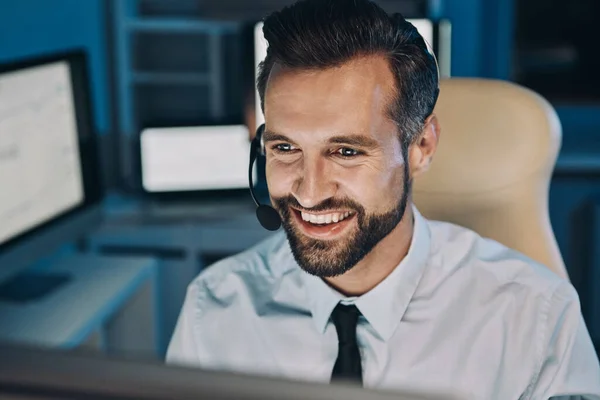 Joven feliz con auriculares mirando el ordenador y sonriendo mientras se queda hasta tarde en la oficina — Foto de Stock