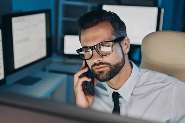 Joven condensado hablando por teléfono móvil y mirando el ordenador mientras se queda hasta tarde en la oficina — Foto de Stock