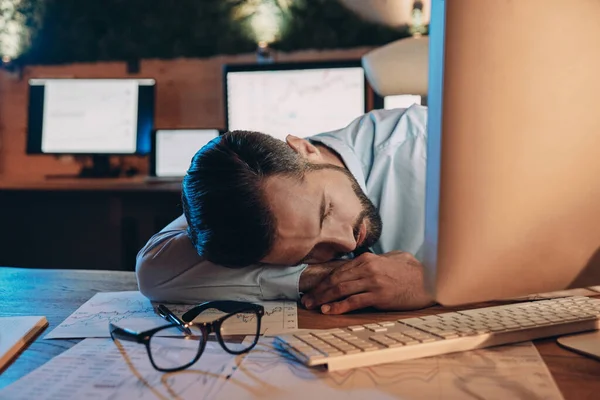 Erschöpfter junger Mann schläft am Arbeitsplatz, während er spät im Büro bleibt — Stockfoto