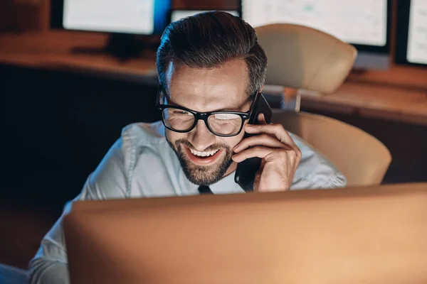 Visão superior do jovem feliz falando no telefone celular e olhando para o computador enquanto permanece até tarde no escritório — Fotografia de Stock