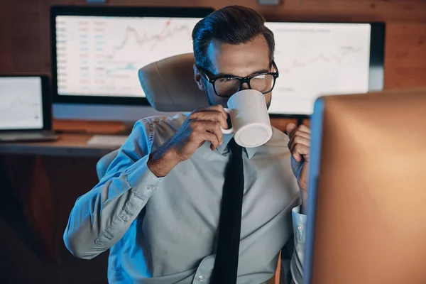Pewny siebie młody człowiek pijący kawę i pracujący na komputerze zostając do późna w biurze — Zdjęcie stockowe