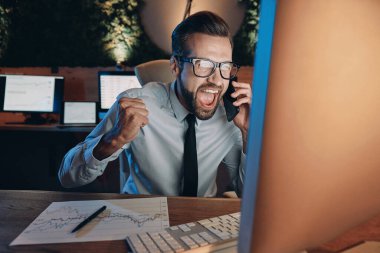 Ofiste geç saatlere kadar kalırken cep telefonuyla konuşan ve el kol hareketi yapan mutlu genç adam.