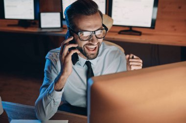 Ofiste geç saatlere kadar kalırken cep telefonuyla konuşan ve el kol hareketi yapan mutlu genç adam.