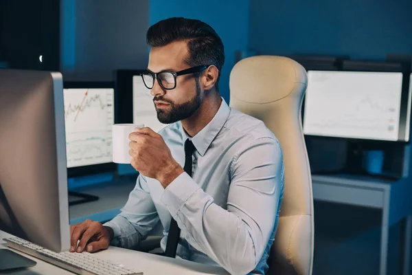 Selbstbewusster junger Mann trinkt Kaffee und arbeitet am Computer, während er spät im Büro bleibt — Stockfoto
