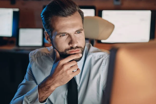 Вдумчивый молодой человек смотрит в компьютер, сидя допоздна в офисе — стоковое фото