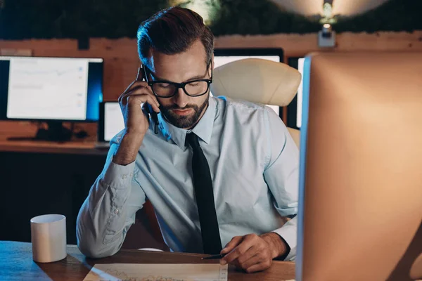 Joven confiado trabajando en la computadora y hablando por teléfono móvil mientras se queda hasta tarde en la oficina — Foto de Stock