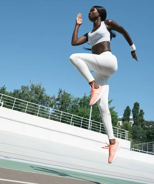 Mulher africana jovem confiante em roupas esportivas pulando na pista ao ar livre — Fotografia de Stock