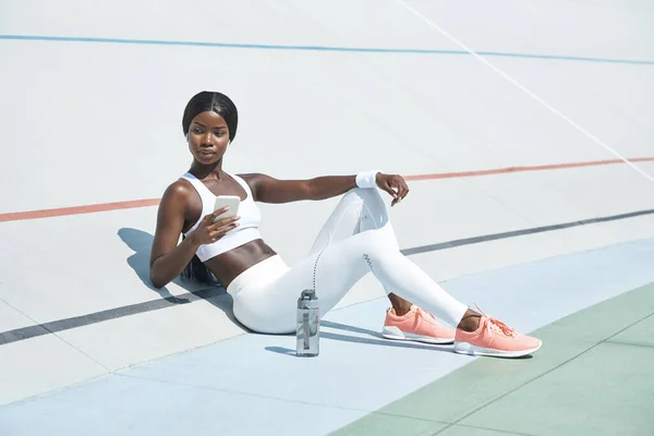 Hermosa mujer africana joven en ropa deportiva sosteniendo el teléfono inteligente mientras está sentado en pista deportiva al aire libre —  Fotos de Stock