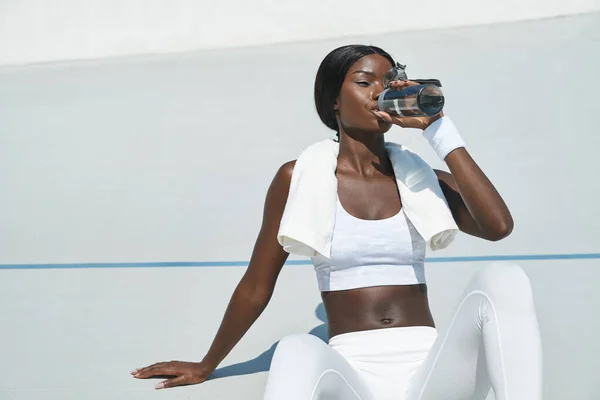 Красивая молодая африканка в спортивной одежде пьет воду, сидя на спортивной трассе на открытом воздухе — стоковое фото