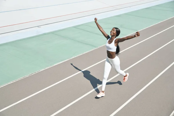 Draufsicht auf eine glückliche junge Afrikanerin in Sportkleidung, die auf der Strecke läuft und die Arme hochreißt — Stockfoto