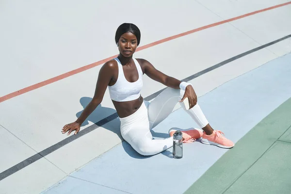 Schöne junge Afrikanerin in Sportkleidung, die eine Flasche mit Wasser hält, während sie im Freien auf einer Sportbahn sitzt — Stockfoto