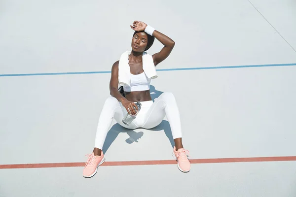 Fatiguée jeune femme africaine en vêtements de sport tenant bouteille avec de l'eau et touchant le front — Photo