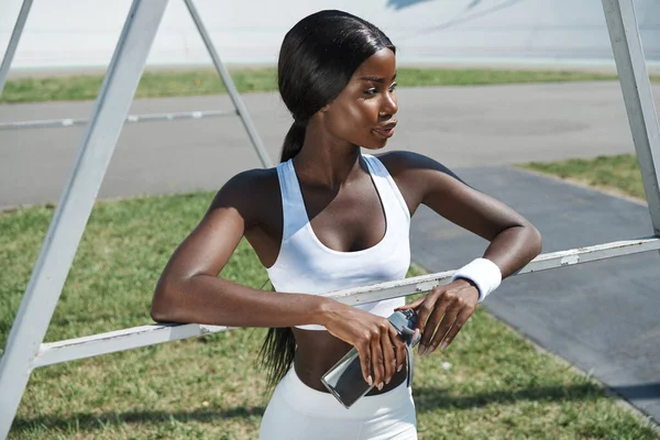 Hermosa joven africana en ropa deportiva sosteniendo botella con agua mientras está de pie al aire libre —  Fotos de Stock