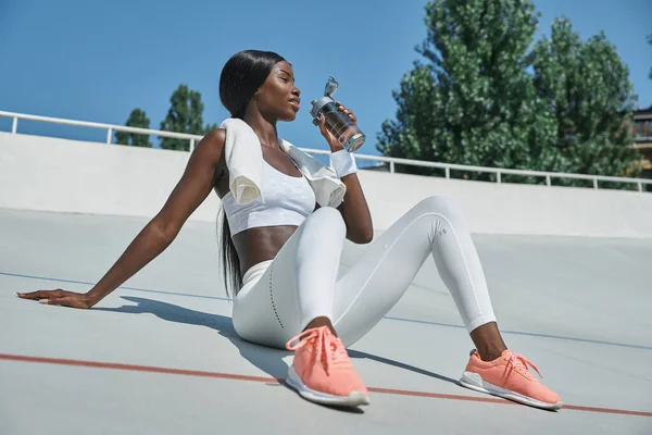 Mooie jonge Afrikaanse vrouw in sportkleding drinkwater tijdens het zitten op sportbaan buiten — Stockfoto