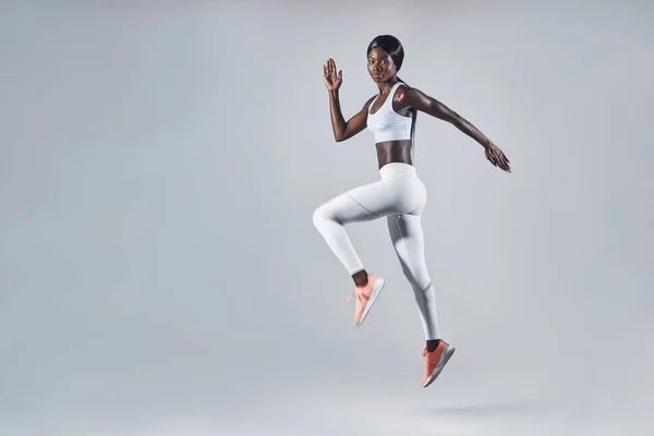Longitud completa de mujer africana joven y confiada en ropa deportiva saltando sobre fondo gris — Foto de Stock