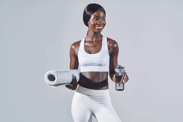 Feliz joven africana en ropa deportiva sosteniendo botella con agua y colchoneta de ejercicio —  Fotos de Stock