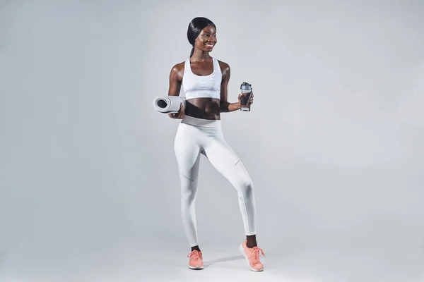 Šťastná mladá africká žena ve sportovním oblečení drží láhev s vodou a cvičení podložka — Stock fotografie