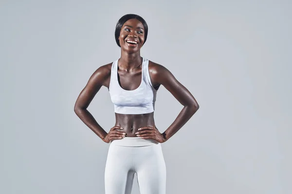 Mujer africana joven y segura en ropa deportiva cogida de la mano en la cadera mientras está de pie sobre un fondo gris —  Fotos de Stock