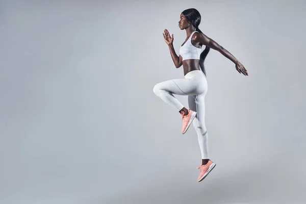 Volledige lengte van zelfverzekerde jonge Afrikaanse vrouw in sportkleding springen tegen grijze achtergrond — Stockfoto