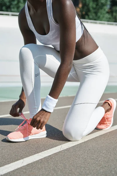 Primer plano de la joven africana vestida con ropa deportiva atando cordones mientras está parada en una pista de atletismo al aire libre —  Fotos de Stock