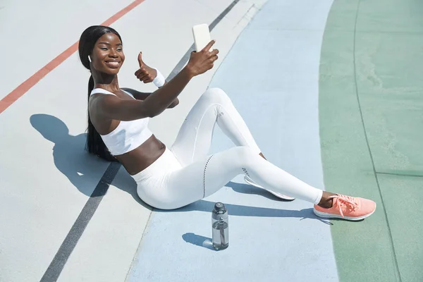 Hermosa joven africana en ropa deportiva teniendo videollamada y sonriendo —  Fotos de Stock