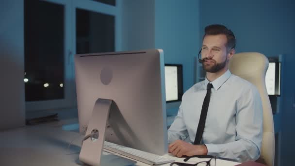 Jovem empresário feliz no fone de ouvido olhando para o computador e sorrindo — Vídeo de Stock